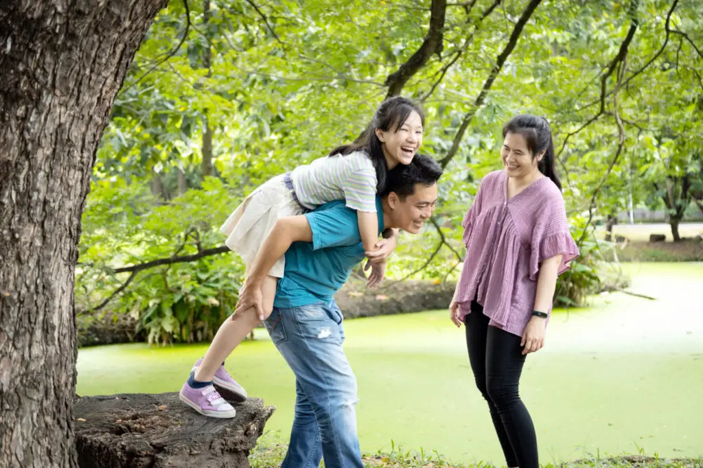 Happy asian family,father laugh giving piggyback ride to beautiful daughter having fun together, smiling mother enjoying active game with family, man carrying child girl on back, play in outdoor park